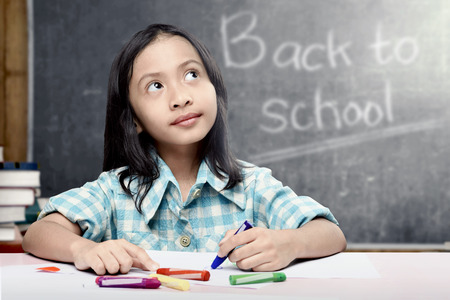 Asian student girl drawing on white paper with colorful crayons in the classroom with piles of books and blackboard background. Back to School conceptの写真素材