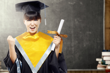 Asian woman in mortarboard hat and diploma graduating from college in the classroom with piles of books and blackboard background. Graduation conceptの写真素材