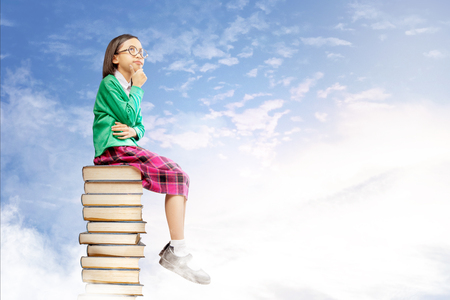 Asian cute girl with glasses think while sitting on the pile of books with blue sky background. Back to School conceptの写真素材