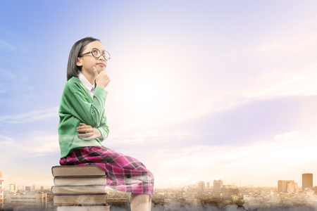 Asian cute girl with glasses think while sitting on the pile of books with city and blue sky background. Back to School conceptの写真素材