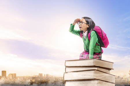 Asian cute girl with glasses and backpack sitting on the pile of books with city and blue sky background. Back to School conceptの写真素材