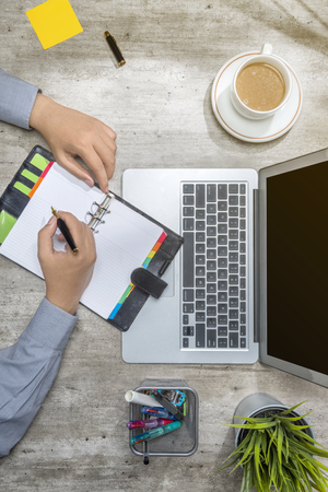 Top view of businessman writing in notebook with laptop, coffee, sticky notes, potted plant and business accessories on the deskの写真素材