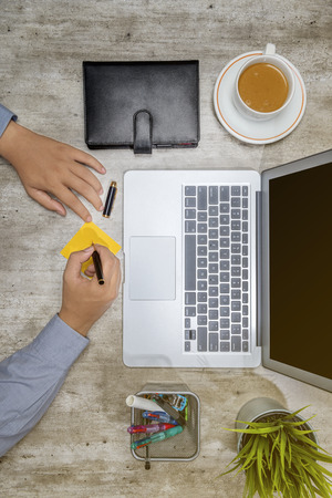 Top view of businessman writing in sticky notes with laptop, coffee, notebook, potted plant and business accessories on the deskの写真素材