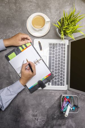 Top view of businessman writing in notepad with laptop, coffee, potted plant and business accessories on the deskの写真素材