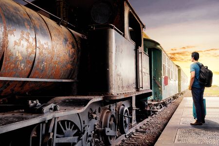 Rear view of asian man with suitcase bag and backpack standing and waiting a train on station with sunset backgroundの写真素材