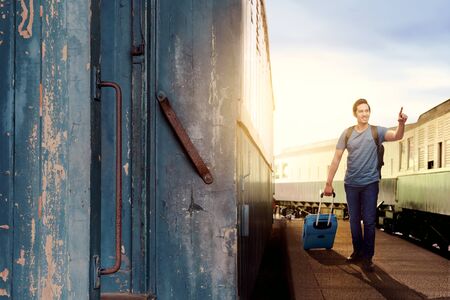 Asian man with suitcase bag and backpack standing and pointing a train wagon on station with blue sky backgroundの写真素材