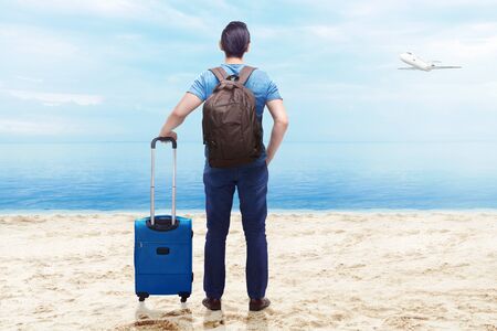 Rear view of asian man with suitcase bag and backpack standing on the beach with flying airplane above the sea and blue sky backgroundの写真素材