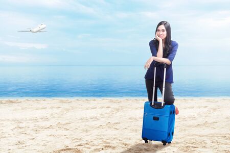 Asian woman with suitcase bag standing on the beach with flying airplane above the sea and blue sky backgroundの写真素材