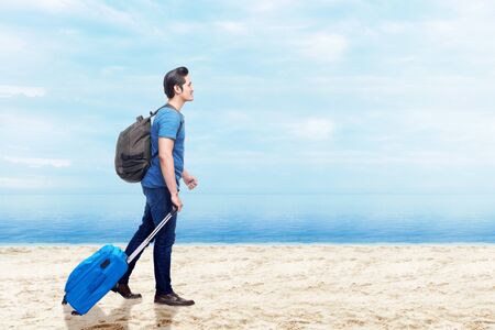 Asian man with suitcase bag and backpack walking on the beach with blue sky backgroundの写真素材
