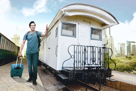 Asian man with suitcase bag and backpack standing and pointing a white train wagon on station with city and blue sky backgroundの写真素材