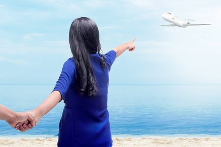 Rear view of asian woman holding her boyfriend hands on the beach and pointing to a flying plane above the sea with blue sky backgroundの写真素材