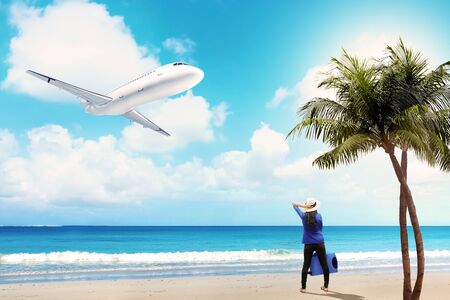 Rear view of asian woman in hat with suitcase bag standing on the beach with flying airplane above the sea and blue sky backgroundの写真素材