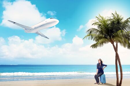 Asian woman in hat sitting on suitcase bag on the beach with flying airplane above the sea and blue sky backgroundの写真素材