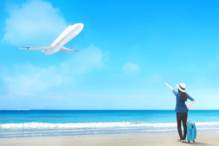 Rear view of asian woman in hat with suitcase bag standing on the beach and pointing to a flying plane above the seaの写真素材