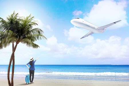 Rear view of asian woman in hat with suitcase bag standing on the beach and pointing to a flying plane above the seaの写真素材