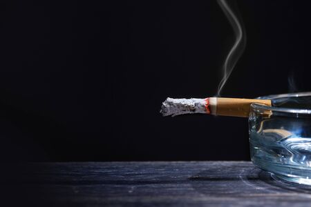 Cigarette on transparent ashtray on wooden table with black background