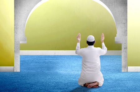 Rear View of Asian Muslim man sitting in pray position while raised hands and praying inside the mosqueの写真素材