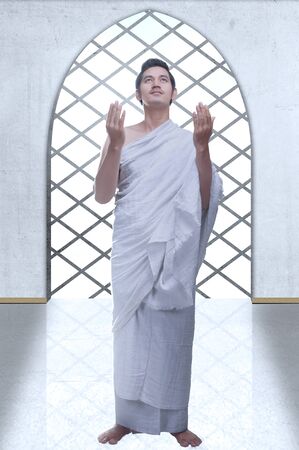 Asian Muslim man in ihram clothes standing and praying while raised arms inside the mosqueの写真素材