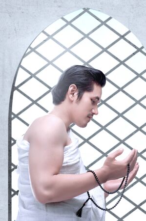 Asian Muslim man in ihram clothes standing and praying with prayer beads while raised arms inside the mosqueの写真素材