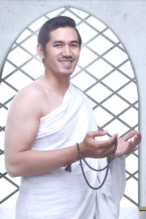 Asian Muslim man in ihram clothes standing and praying with prayer beads while raised arms inside the mosqueの写真素材