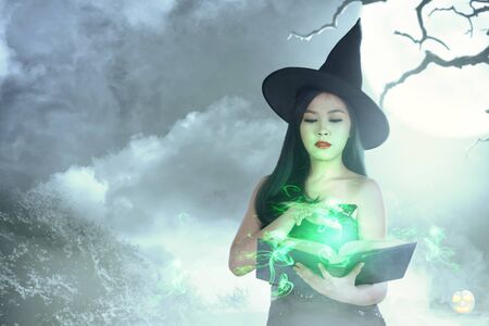 Asian witch woman in hat learns the spell from the magic book with moonlight background. Halloween conceptの写真素材