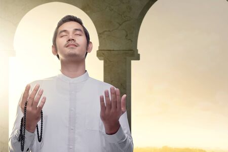Asian Muslim man standing and praying with prayer beads while raised arms inside the mosque