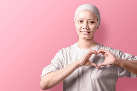Asian woman in a white shirt showing a heart sign with her hands over pink background. Breast cancer...