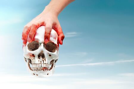 Hand with blood holding a human skull with a sky background