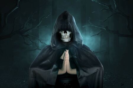 Witch with human skull head in black hooded doing ritual magic on the dark forest