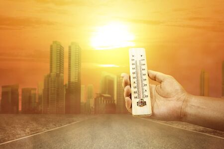 Hand holding thermometer with high temperature on the city with glowing sun background. Heatwave conceptの写真素材