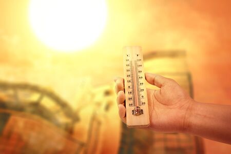 Hand holding thermometer with high temperature on the city with glowing sun background. Heatwave conceptの写真素材