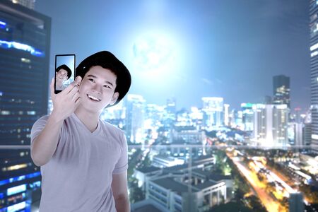 Asian man in hat making selfie using the camera phone with skyscrapers background at night