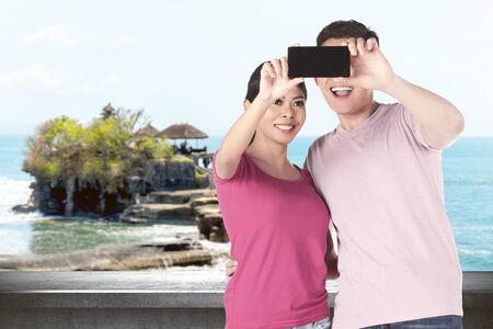 Asian couples making a selfie using a camera phone with Tanah Lot Temple backgroundの写真素材