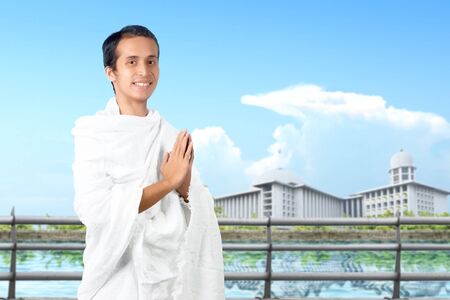 Asian Muslim man in ihram clothes standing and praying with mosque view background