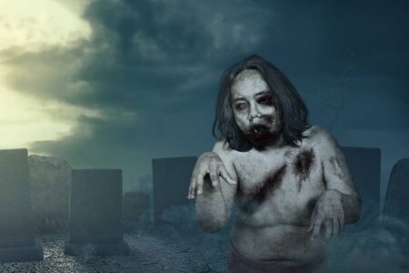 Scary zombie with blood and wound on his body walking around on the graveyard