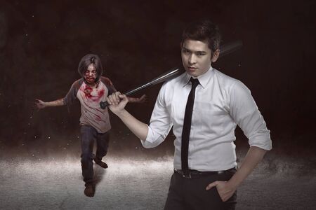 Asian man standing with a baseball bat on his hand facing zombies on the dark roomの写真素材