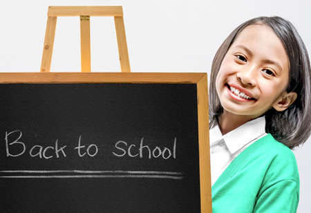 Asian little girl standing with a small chalkboard on a wooden easel inside the classroom. Back to...