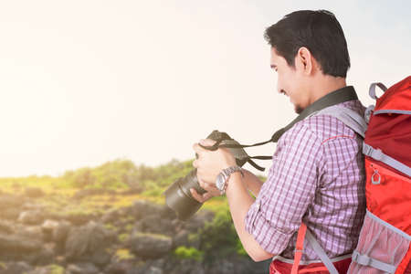 Asian man with a backpack holding camera with a lake view and blue sky background. World Tourism Day
