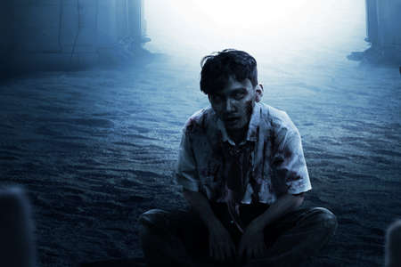 Scary zombie with blood and wound on his body sitting on the graveyardの写真素材