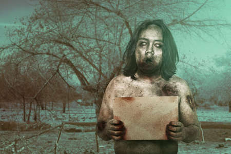 Scary zombie with blood and wound on his body holding an empty board on the field. Empty board for...