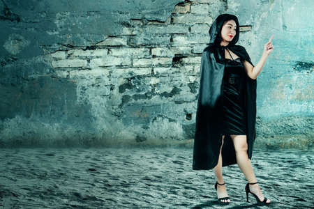 Asian witch woman with a cloak pointing something with a cracked wall background