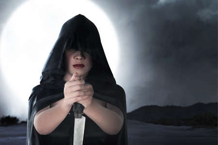 Asian witch woman with a cloak holding a knife standing with night scene backgroundの写真素材