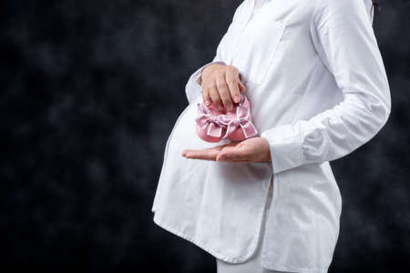 Pregnant woman holding baby girl shoes on her belly with black background