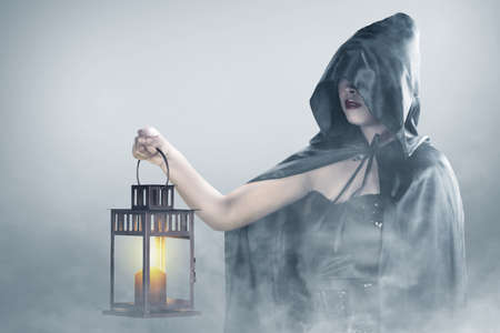 Asian witch woman with a cloak holding a lantern standing in the fogの写真素材