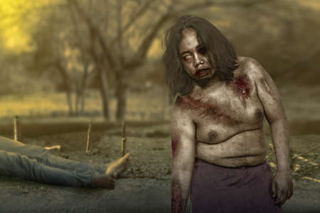 Scary zombie with blood and wound on his body with a dead man on the fieldの写真素材
