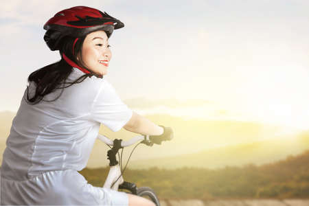 Asian woman with a bicycle helmet riding a bicycle on a wooden path with sunlight backgroundの写真素材