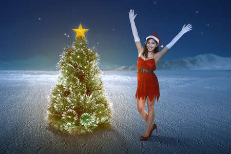 Asian woman in Santa costume standing with a Christmas tree with a night scene background. Merry Christmasの写真素材