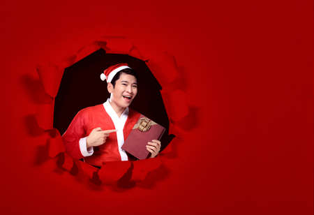 Asian man in Santa costume standing and holding the gift box with a red backgroundの写真素材