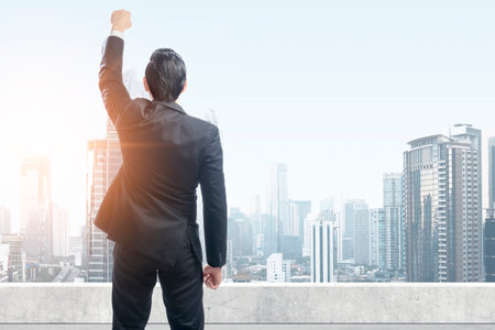 Rear view of Asian businessman standing with raised hand on the rooftop with skyscrapers backgroundの写真素材