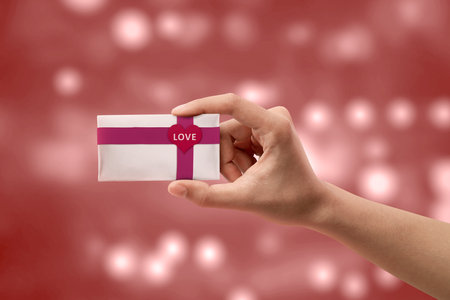 Human hand holding white gift box with red ribbon and heart shape with LOVE text with blurred light background. Valentines dayの写真素材